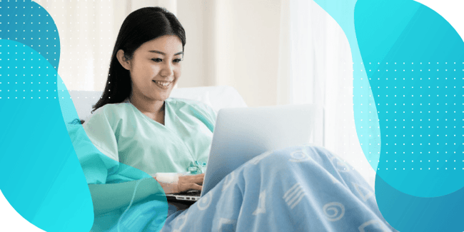 A smiling patient in a hospital gown uses a laptop while sitting in bed, researching the optimization of cancer trials with cognitive assessments.