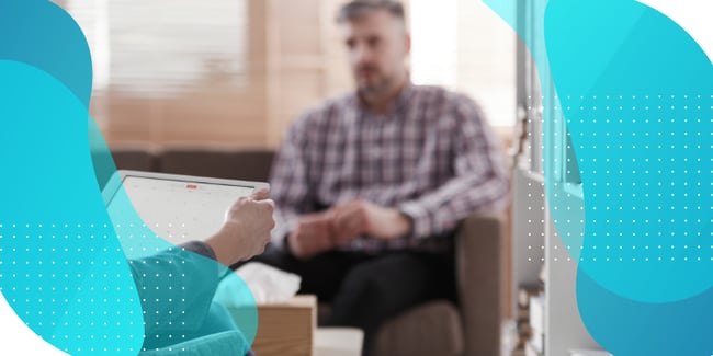 A clinician holds a tablet while a patient sits in the background during a consultation